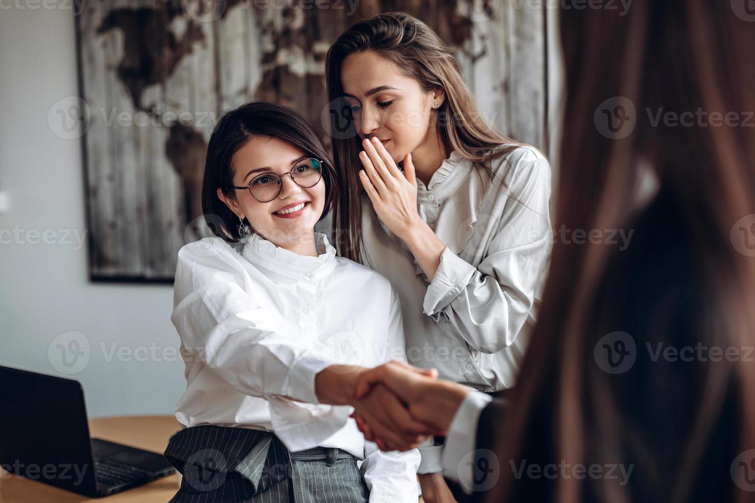 menina sorridente de óculos aperta a mão de seu colega enquanto a assistente diz algo em seu ouvido foto