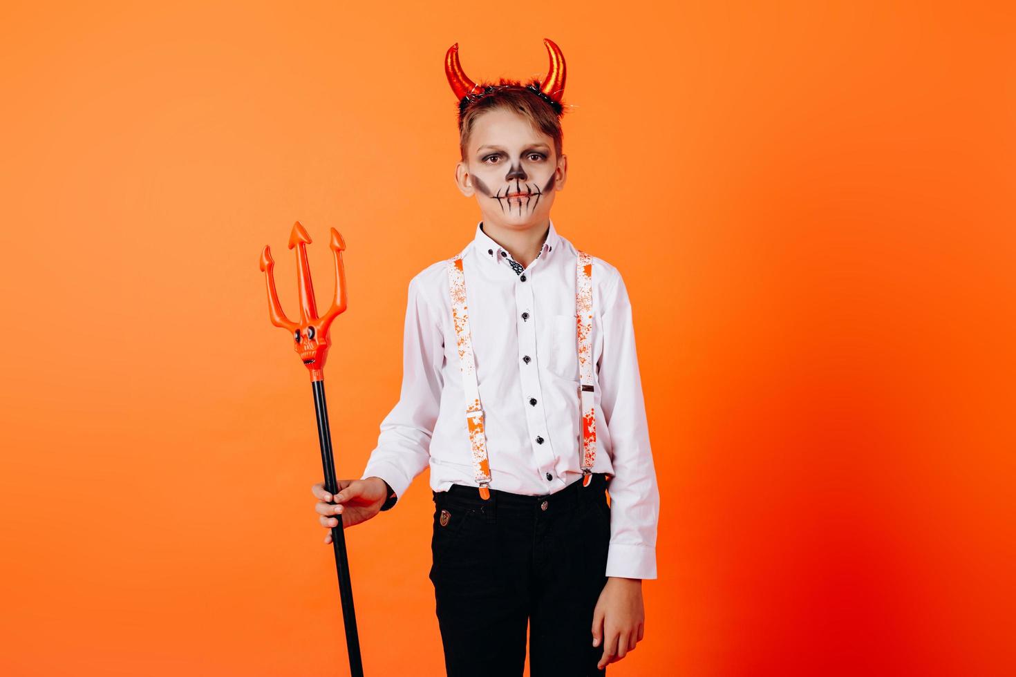 menino diabo em pé contra um fundo laranja em maquiagem de baile de máscaras. conceito de halloween foto