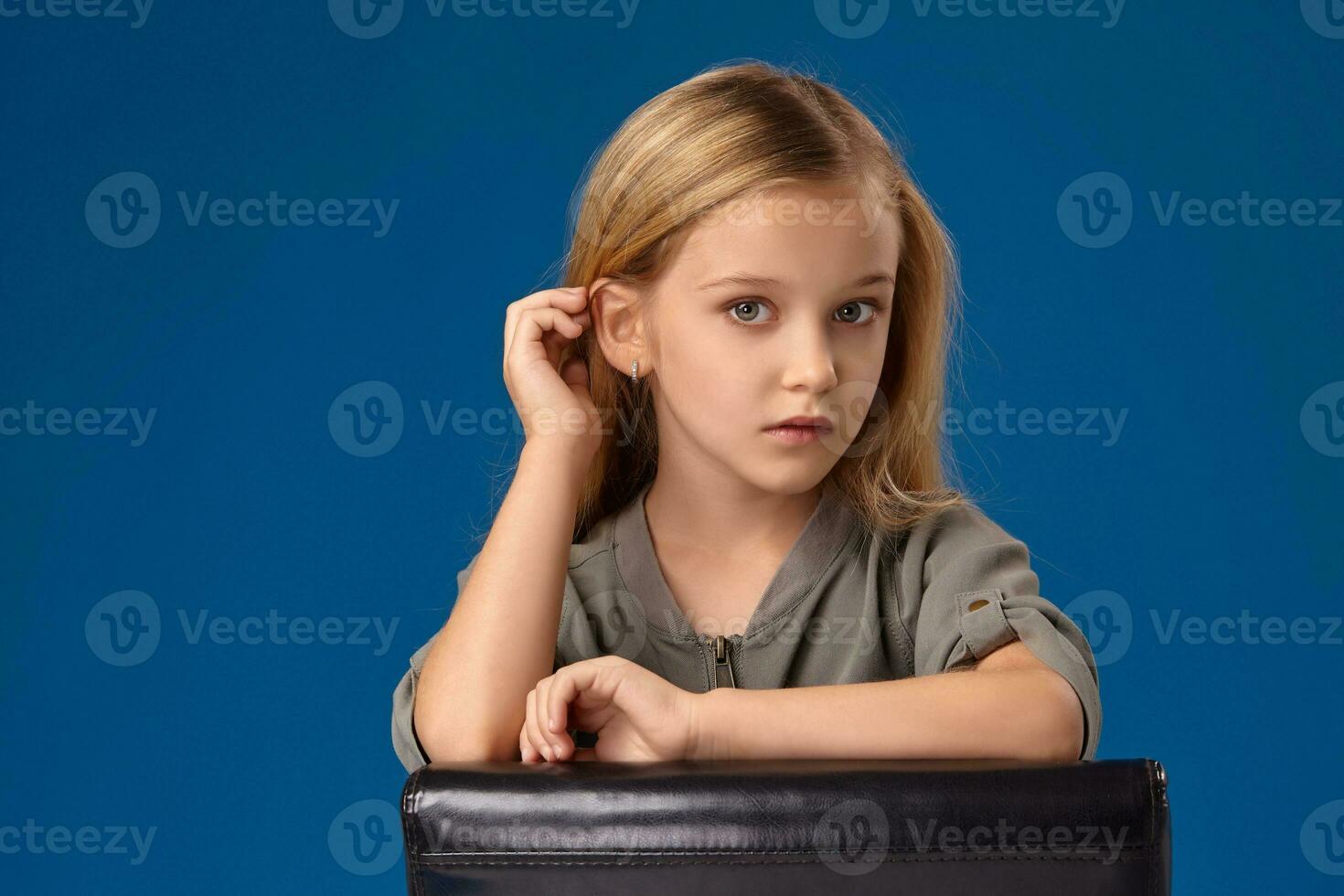 pequeno menina com cinzento olhos e loiro cabelo senta em uma cadeira foto