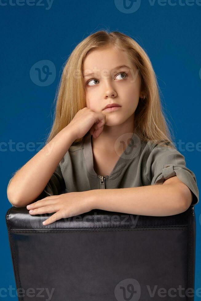 pequeno menina com cinzento olhos e loiro cabelo senta em uma cadeira foto