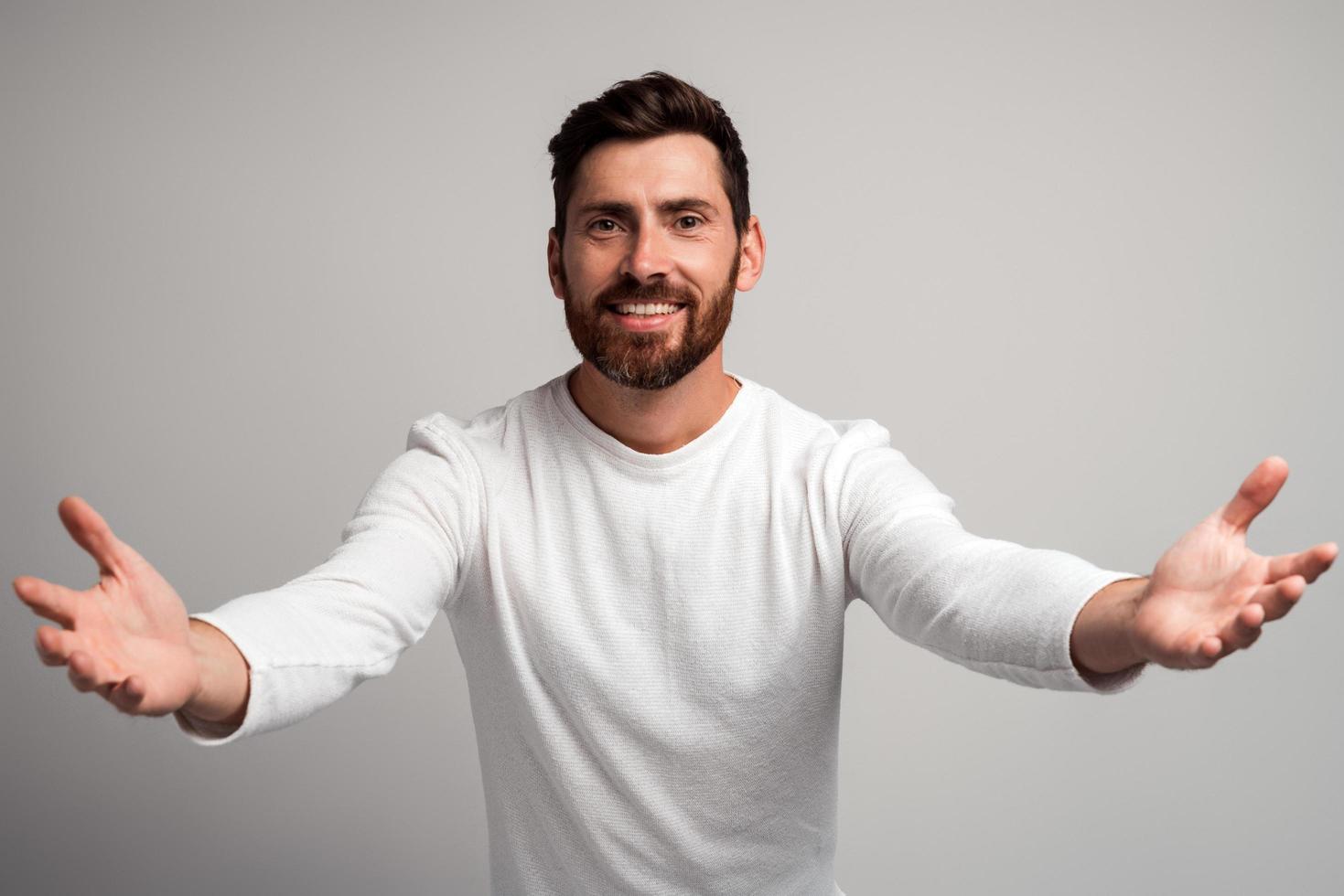 homem generoso de coração aberto com barba, compartilhando abrindo as mãos, olhando para a câmera com um sorriso gentil, saudação e regalando, feliz e feliz em vê-lo. estúdio interno tiro isolado no fundo branco foto