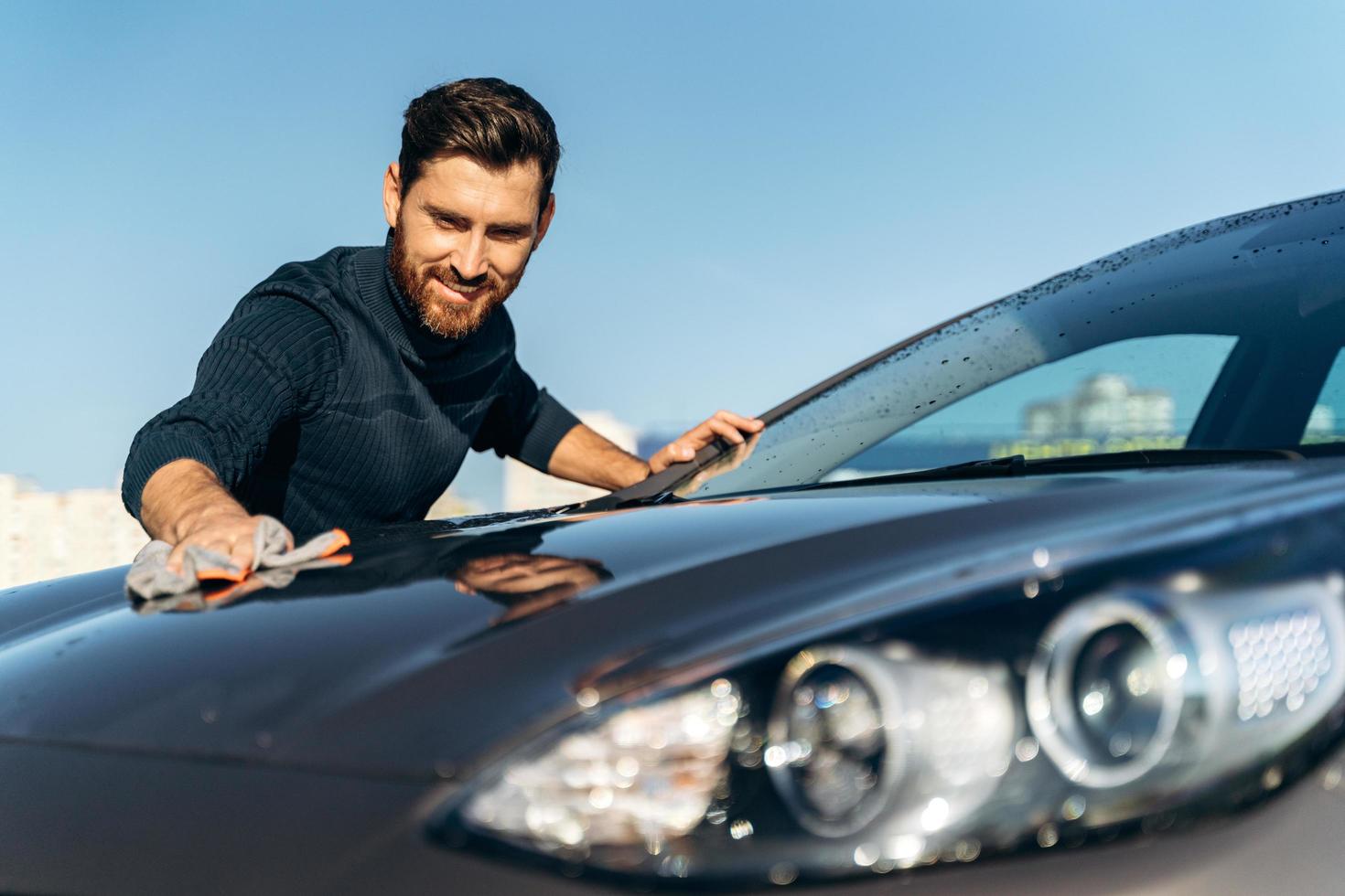 cintura para cima vista do retrato do jovem proprietário do sexo masculino feliz, limpando seu carro preto com um pano em pé na rua durante o dia ensolarado. foto