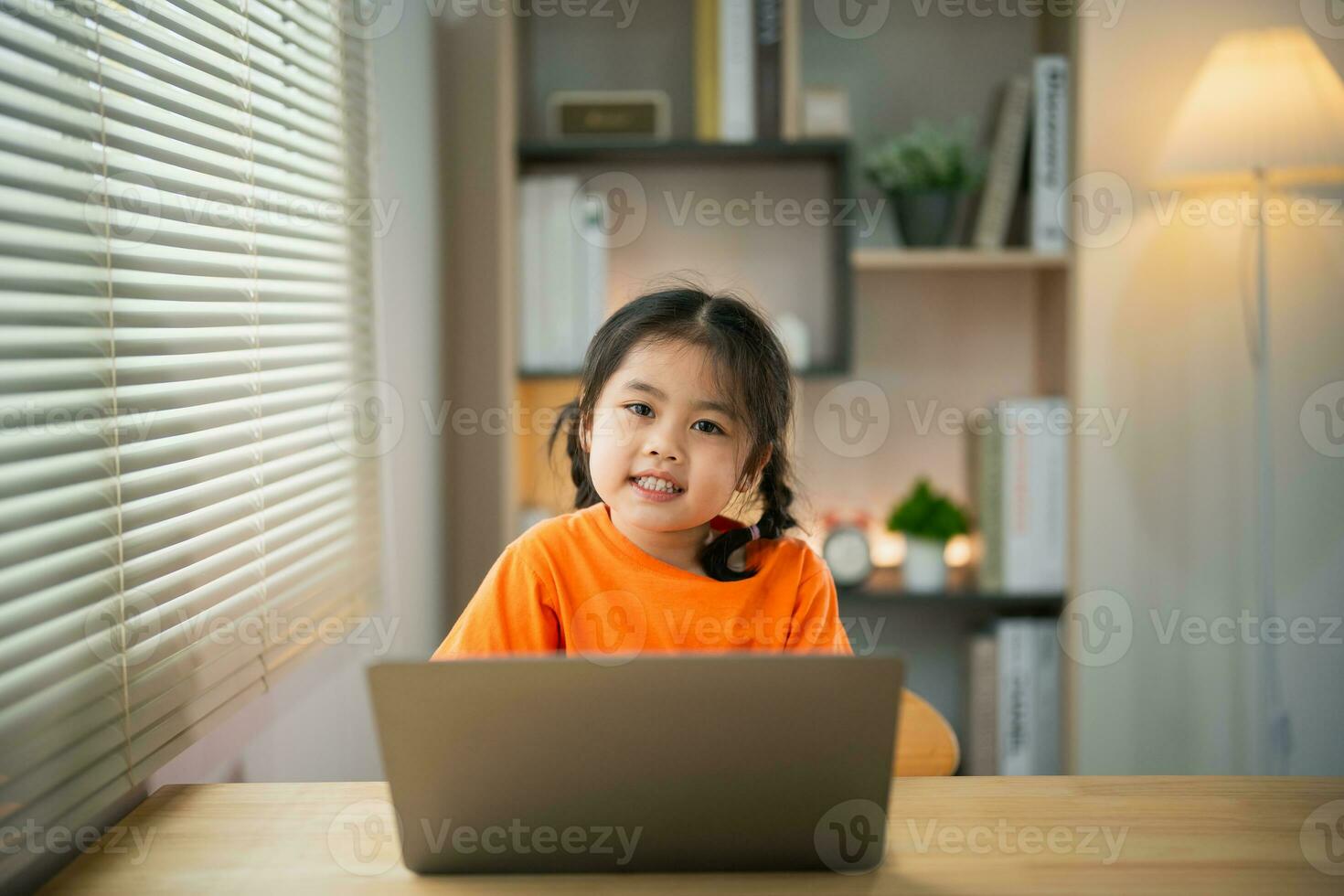 ásia bebê menina vestindo camiseta sorridente concentrado para usar computador portátil e estude conectados em madeira mesa escrivaninha dentro vivo quarto às casa às noite. Educação Aprendendo conectados a partir de casa conceito. foto