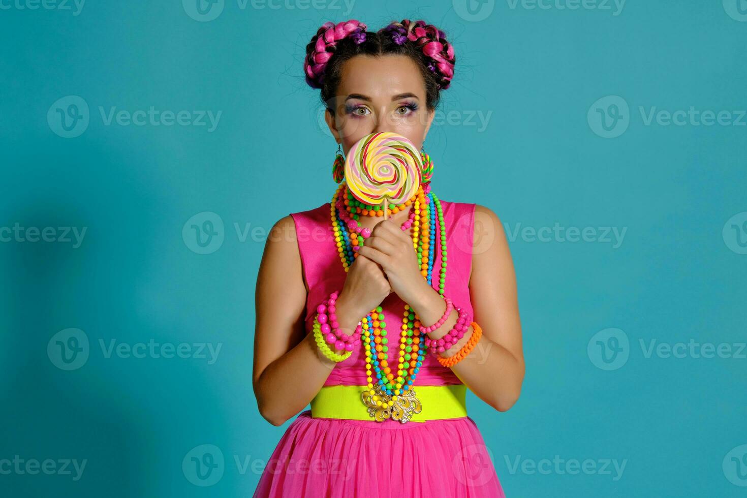 adorável menina com uma multicolorido tranças Penteado e brilhante inventar, posando dentro estúdio contra uma azul fundo, segurando uma pirulito dentro dela mão. foto