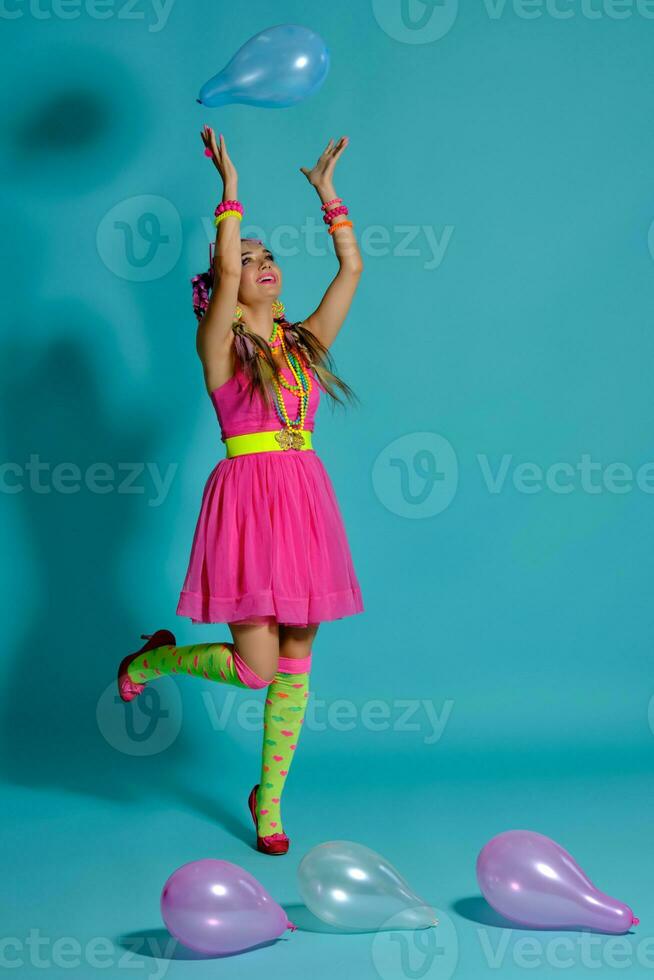 adorável menina com uma multicolorido tranças Penteado e brilhante inventar, posando dentro estúdio com ar balões contra uma azul fundo. foto