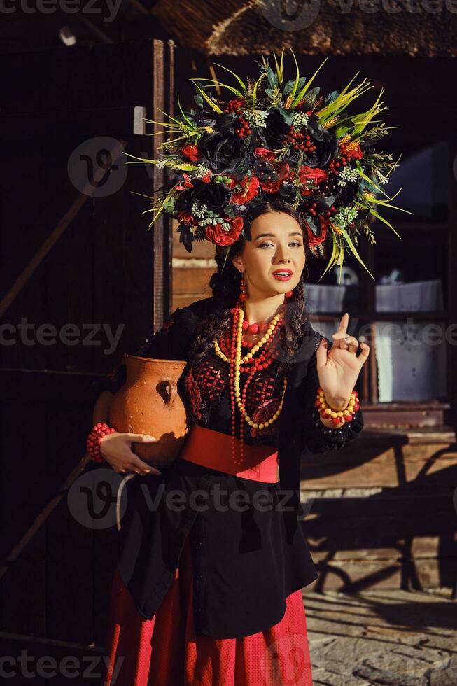 morena menina dentro uma Preto e vermelho ucraniano bordado autêntico nacional traje e uma guirlanda do flores é posando em pé às a portão. foto