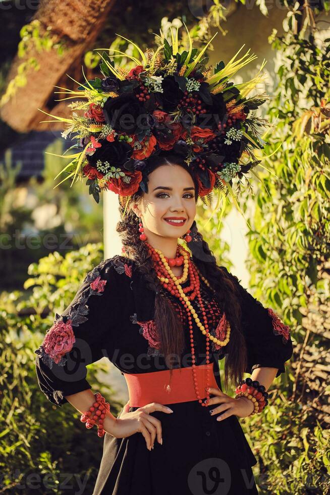 morena menina dentro Preto e vermelho bordado ucraniano autêntico nacional traje e uma guirlanda do flores é posando em pé contra uma branco cabana. foto