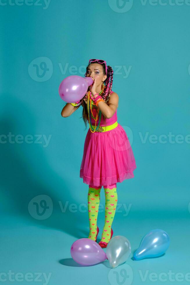 adorável menina com uma multicolorido tranças Penteado e brilhante inventar, posando dentro estúdio com ar balões contra uma azul fundo. foto