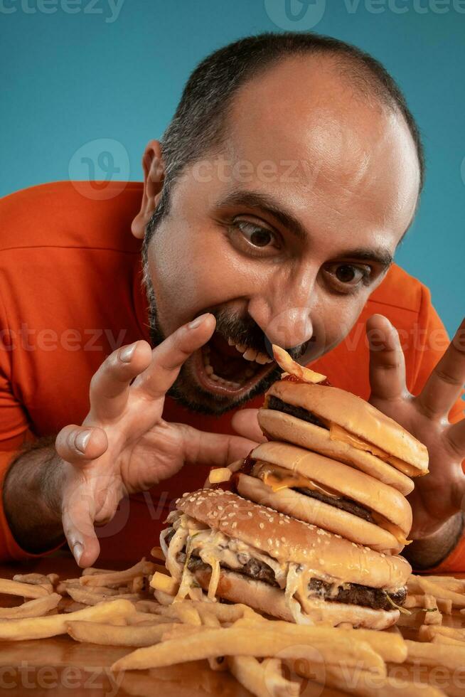 fechar-se retrato do uma meia idade homem com barba, vestido dentro uma vermelho gola alta, posando com hambúrgueres e francês fritas. azul fundo. velozes Comida. foto