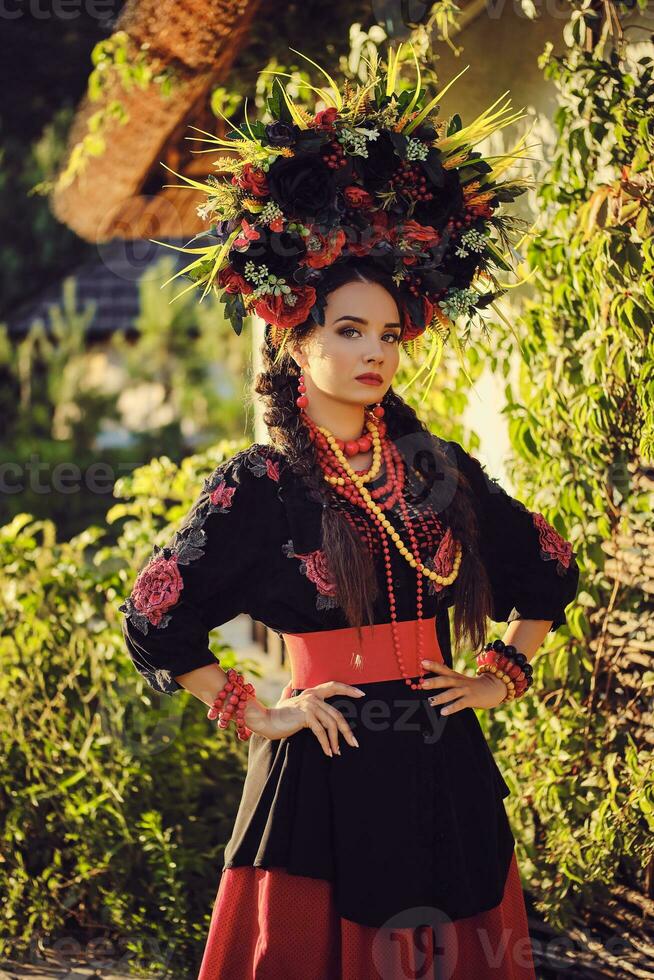 morena menina dentro Preto e vermelho bordado ucraniano autêntico nacional traje e uma guirlanda do flores é posando em pé contra uma branco cabana. foto