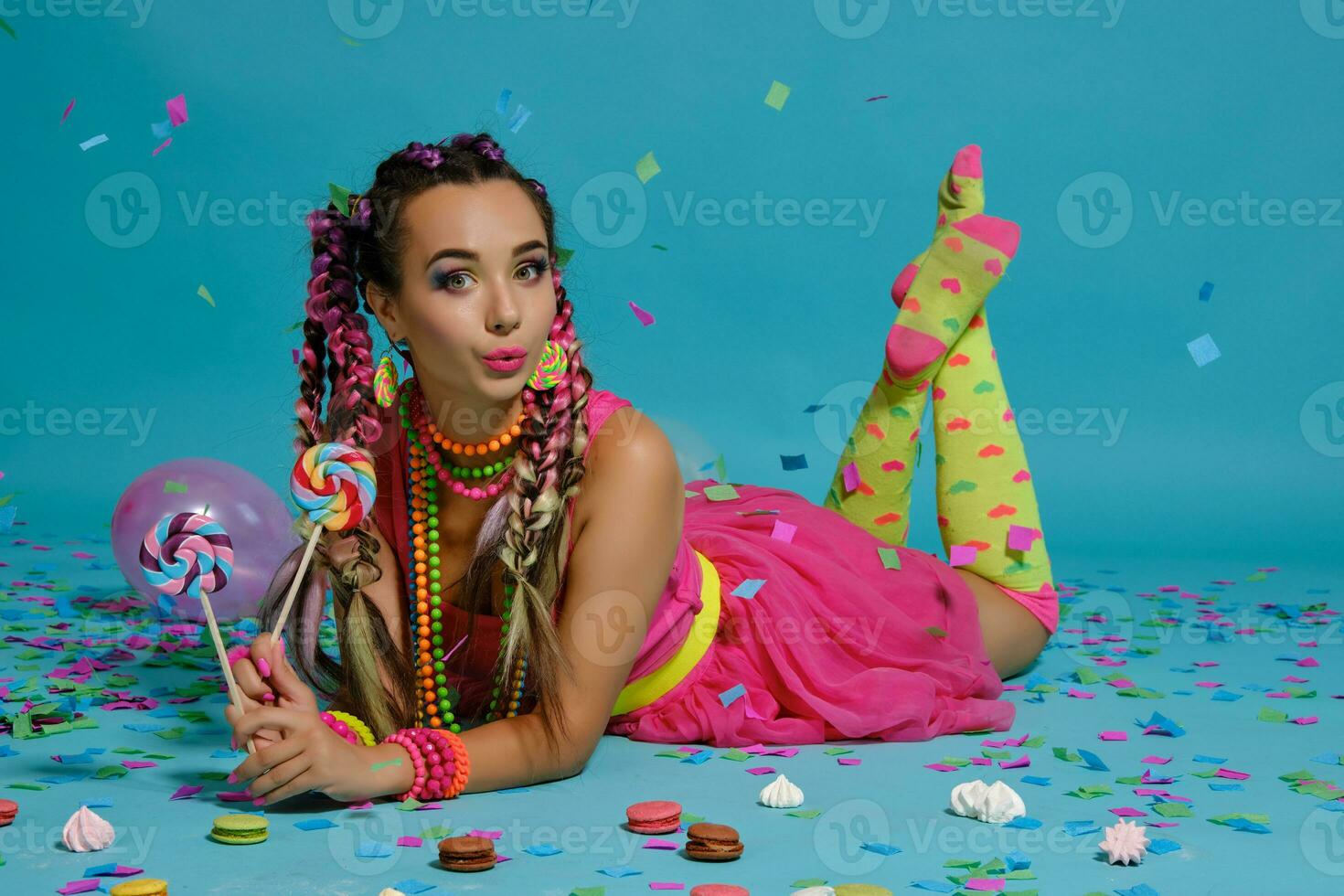 adorável menina com uma multicolorido tranças Penteado e brilhante inventar, posando dentro estúdio com pirulito, ar balões e confete contra uma azul fundo. foto