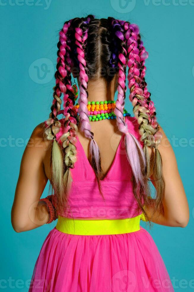 adorável menina com uma multicolorido tranças Penteado e brilhante inventar, posando dentro estúdio contra uma azul fundo. foto