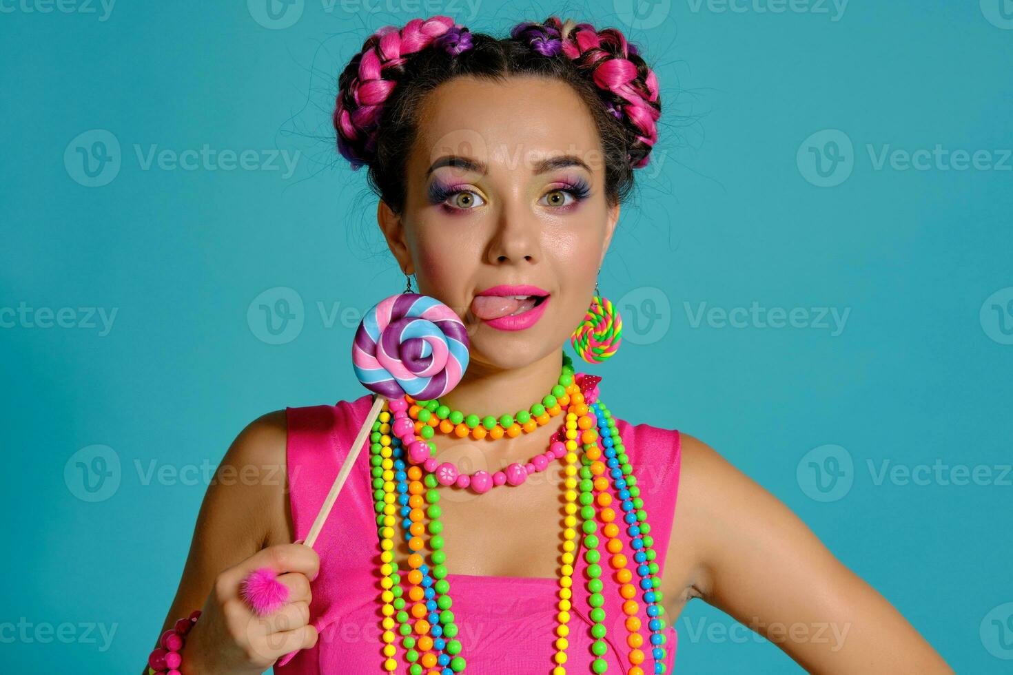 adorável menina com uma multicolorido tranças Penteado e brilhante inventar, posando dentro estúdio contra uma azul fundo, segurando uma pirulito dentro dela mão. foto