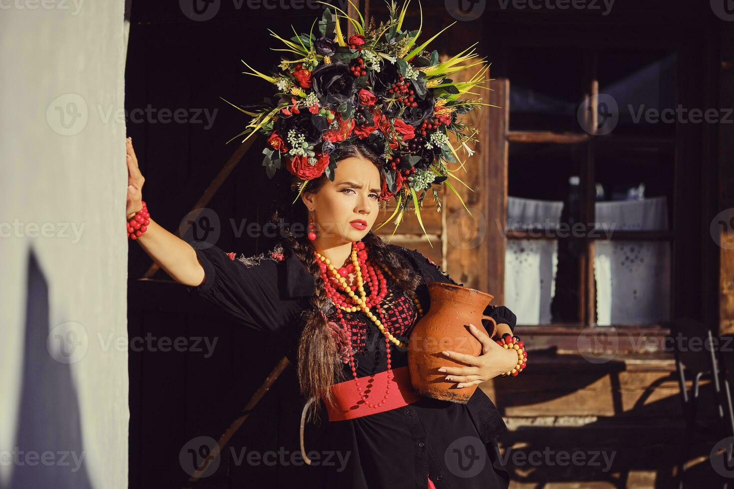 morena menina dentro uma Preto e vermelho ucraniano bordado autêntico nacional traje e uma guirlanda do flores é posando em pé às a portão. foto