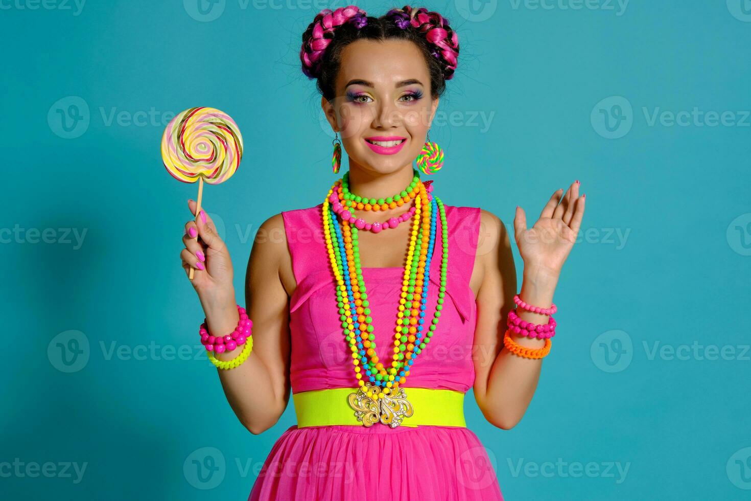 adorável menina com uma multicolorido tranças Penteado e brilhante inventar, posando dentro estúdio contra uma azul fundo, segurando uma pirulito dentro dela mão. foto