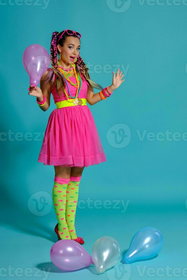 adorável menina com uma multicolorido tranças Penteado e brilhante inventar, posando dentro estúdio com ar balões contra uma azul fundo. foto