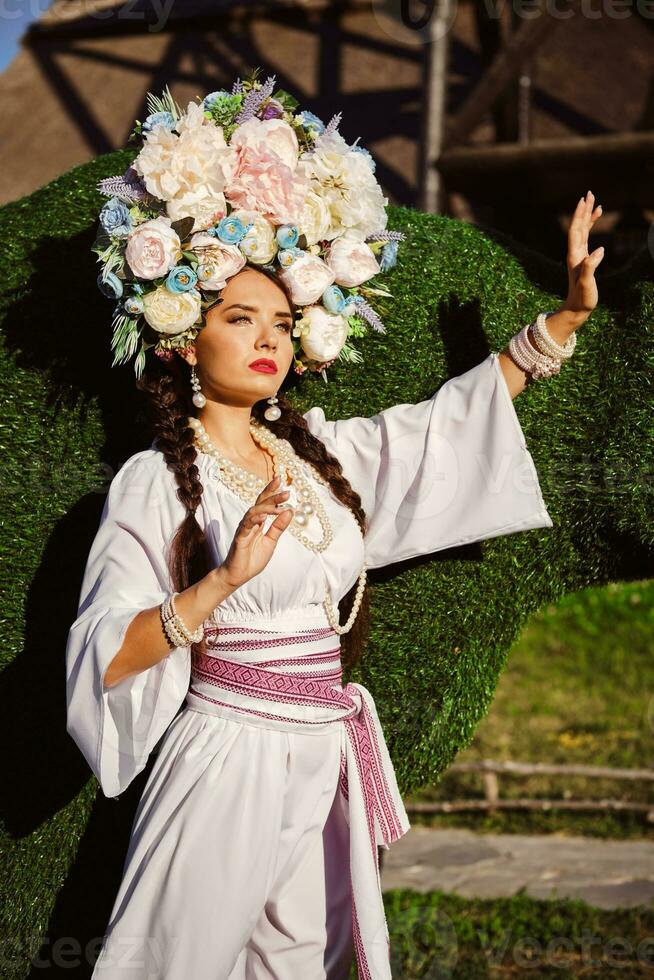 morena menina dentro uma branco ucraniano autêntico nacional traje e uma guirlanda do flores é posando contra uma verde boi esculpido a partir de uma arbusto. foto