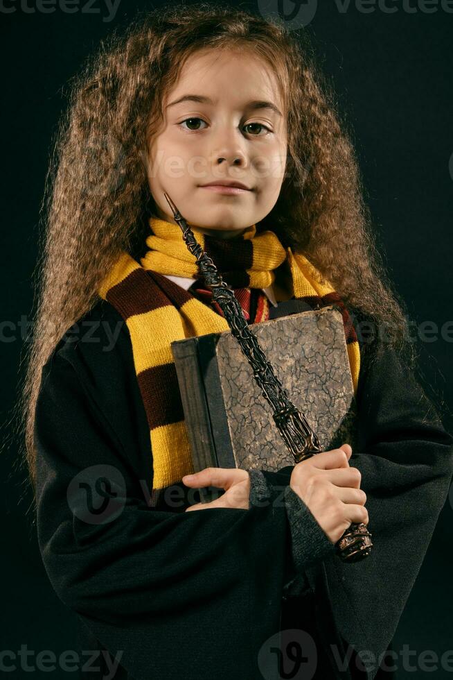 retrato do uma pequeno bruxa menina com grandes Castanho cabelo vestido dentro Sombrio casaco, segurando Magia varinha dentro dela mão, posando em Preto estúdio fundo. foto