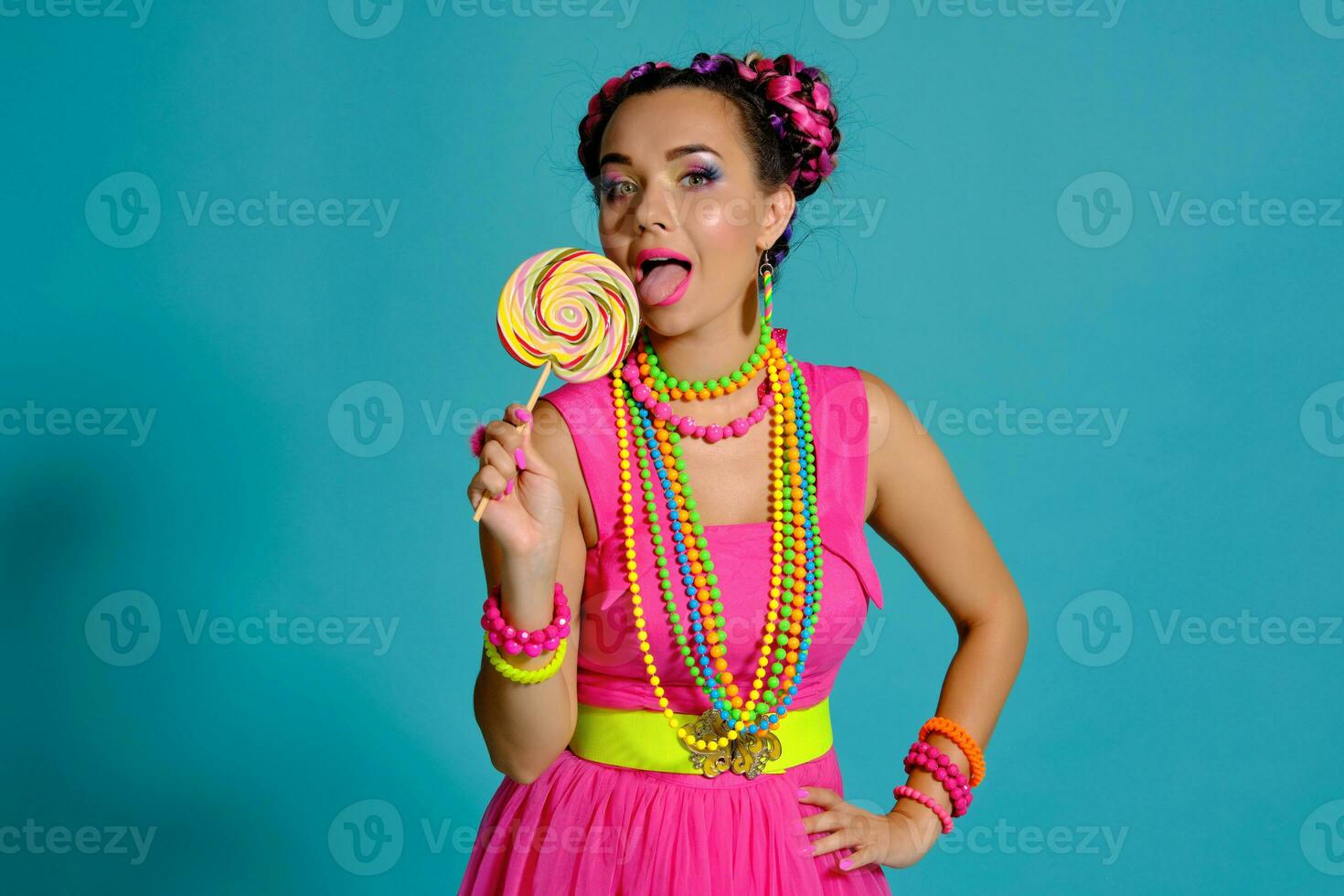 adorável menina com uma multicolorido tranças Penteado e brilhante inventar, posando dentro estúdio contra uma azul fundo, segurando uma pirulito dentro dela mão. foto