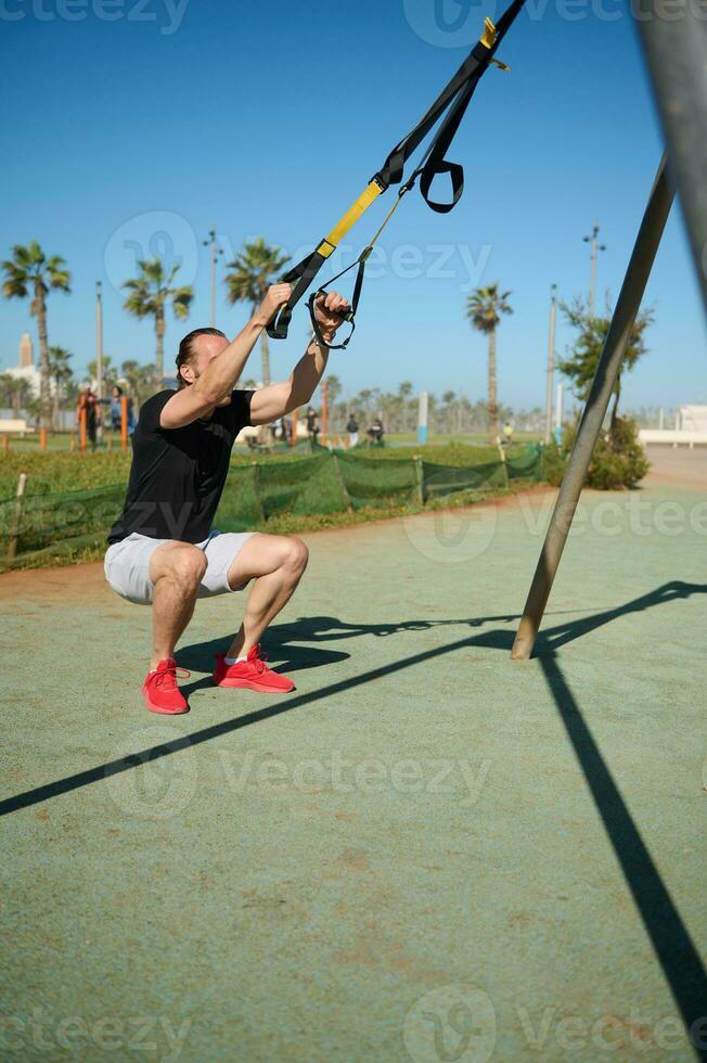 jovem adulto desportista dentro Preto camiseta e cinzento calção exercício com suspensão correias dentro a urbano Esportes terra foto