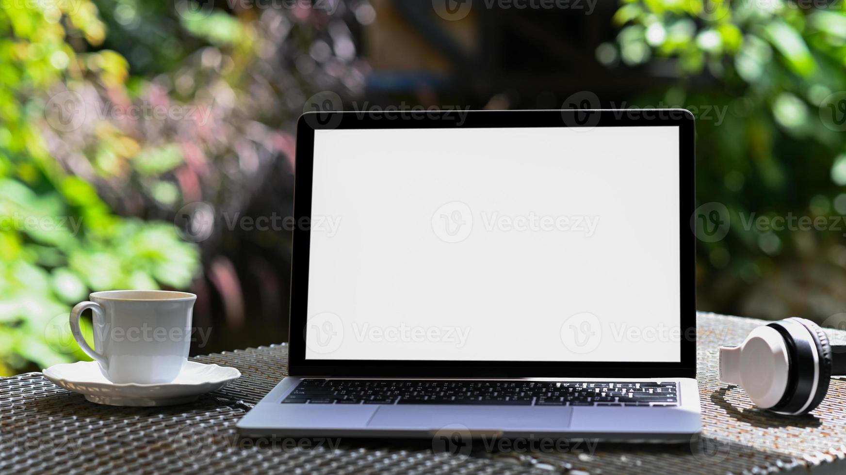 tela em branco do laptop maquete e caneca de café com fone de ouvido na mesa de ferro, fundo verde da árvore. foto