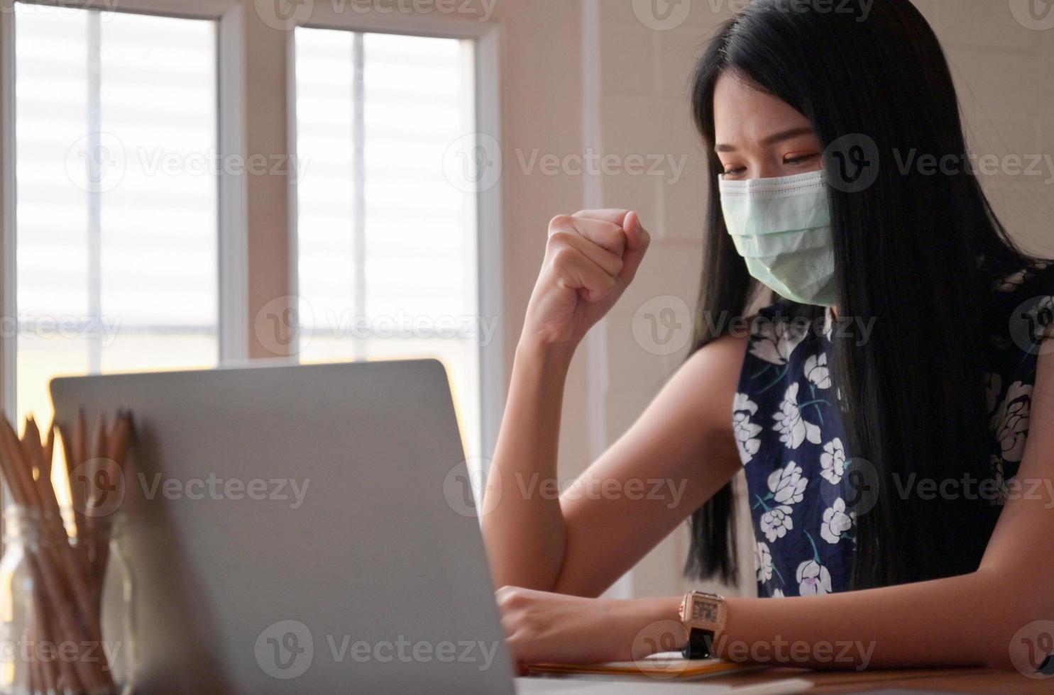 mulher com máscara médica, levantando o punho com o laptop na mesa enquanto trabalhava em casa, trabalha em casa. foto