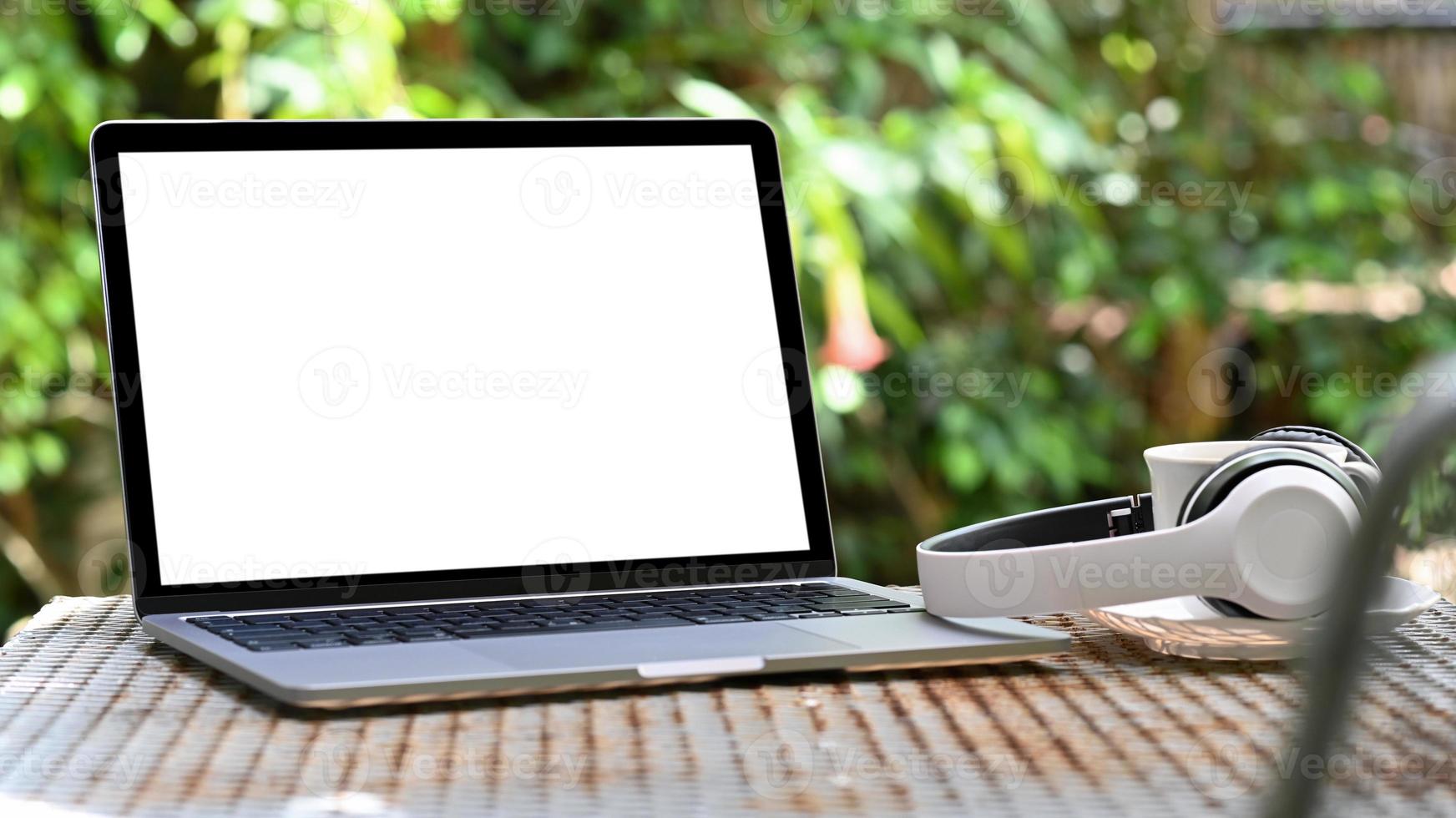 tela em branco do laptop maquete e fone de ouvido com a caneca de café na mesa de ferro, fundo verde da árvore. foto