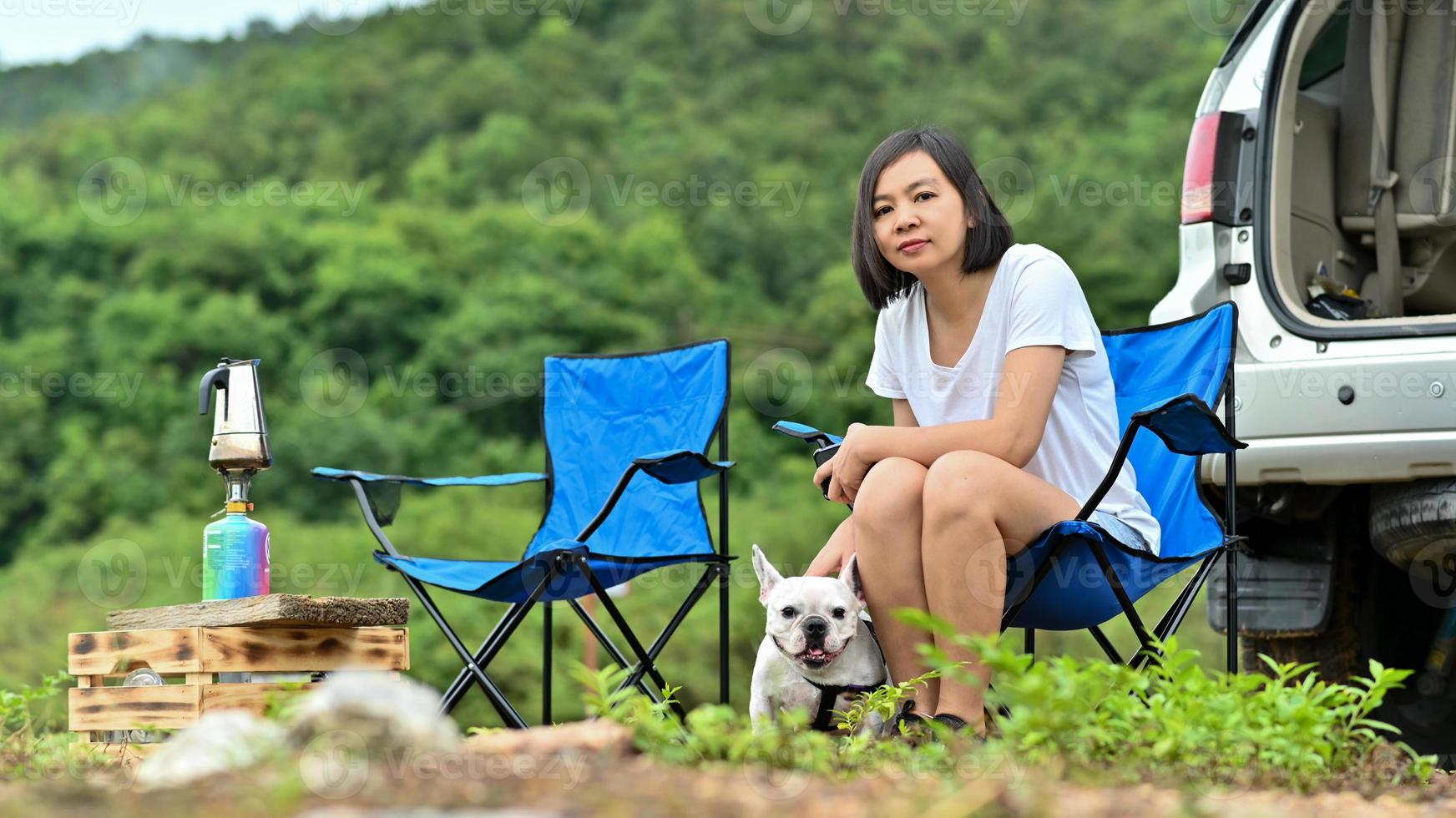 uma mulher em uma camiseta branca e um buldogue francês está relaxando em uma cadeira de camping. foto