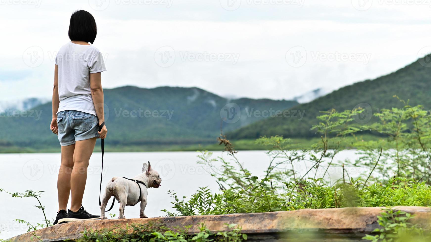 uma mulher em uma camiseta branca e um buldogue francês está de frente para a margem do rio. foto