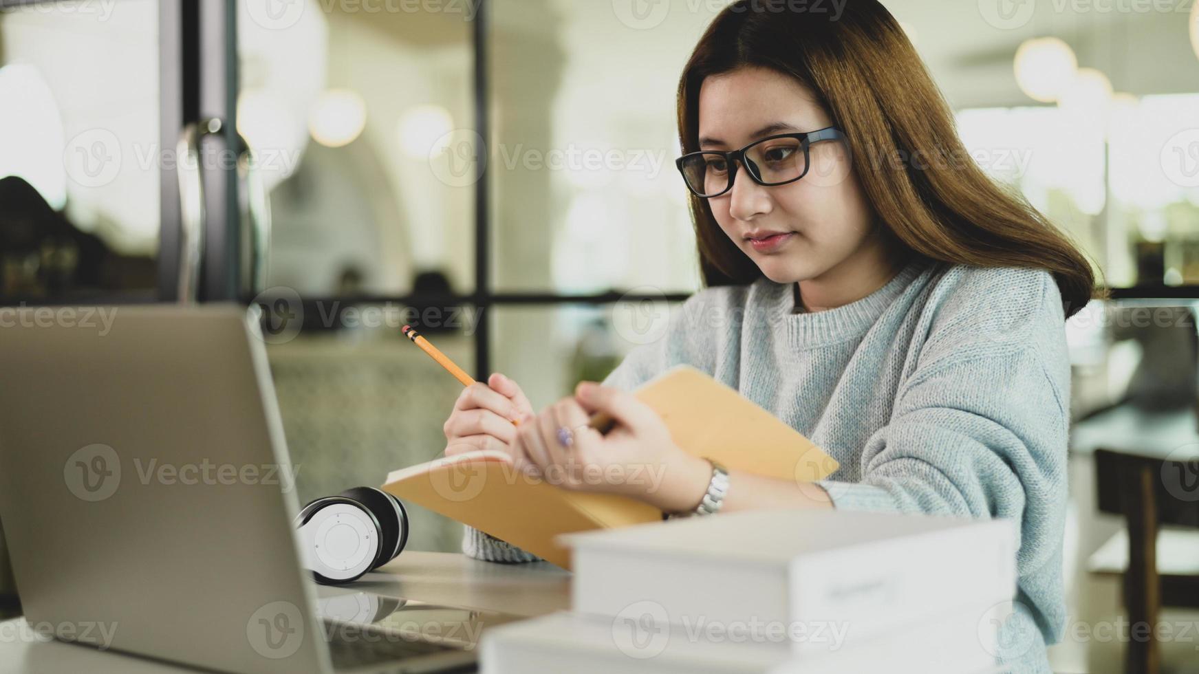 aluna asiática de óculos olhando para o laptop e fazendo anotações em um caderno na mão foto