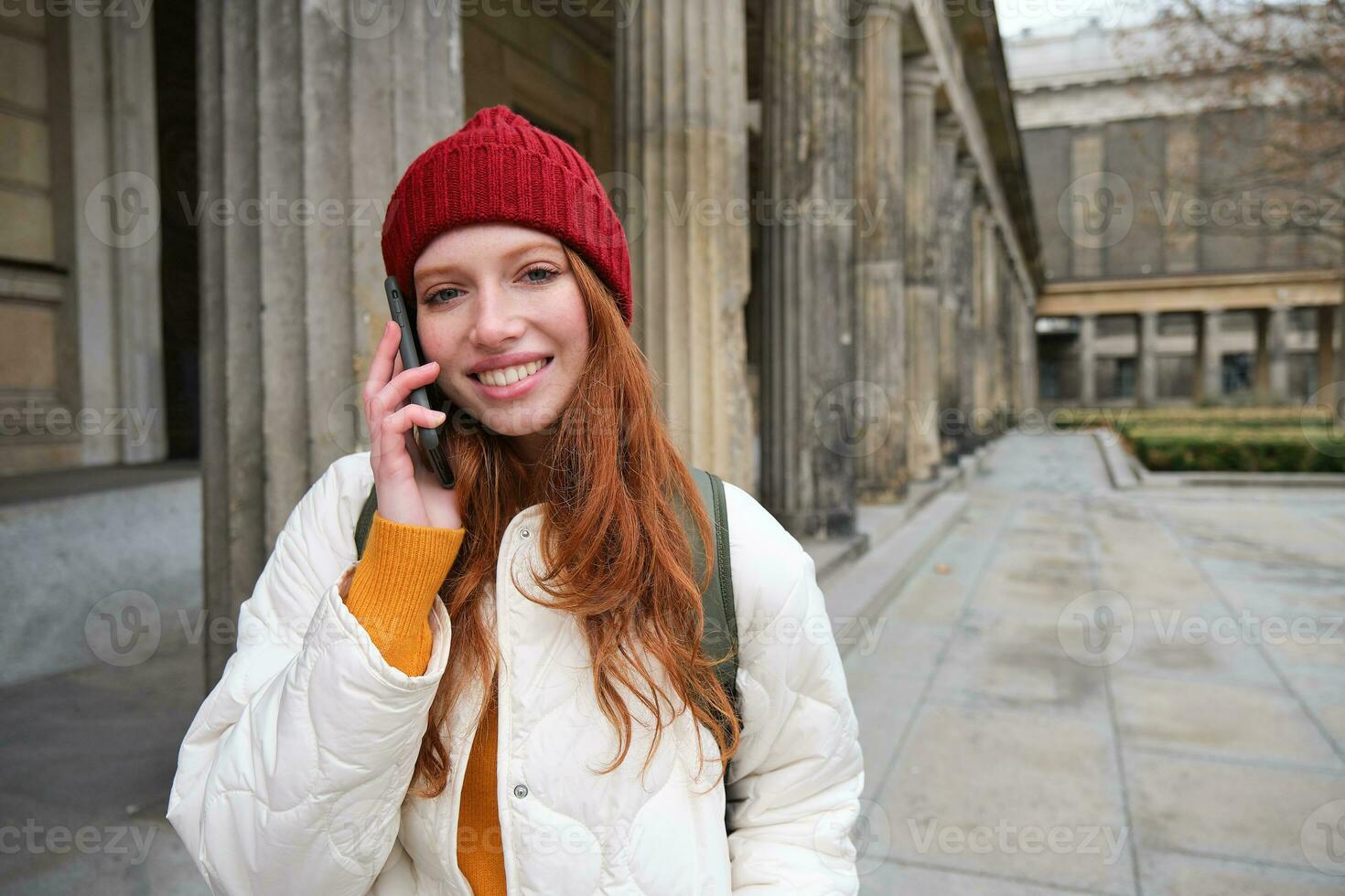 sorridente ruiva fêmea turista fala em Móvel telefone e anda em por aí cidade. feliz aluna dentro vermelho chapéu chamadas amigo, carrinhos em rua e usa Smartphone foto