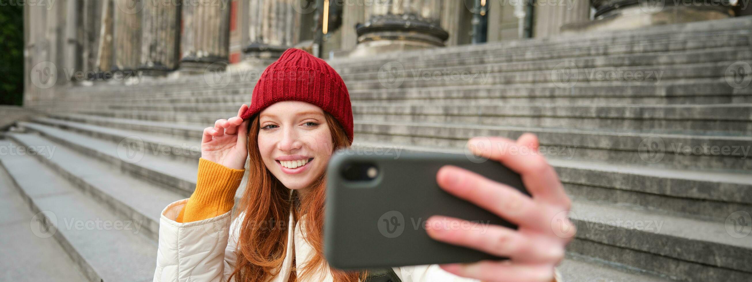 jovem ruiva turista leva selfie dentro frente do museu em escadaria, detém Smartphone e parece às Móvel Câmera, faz foto do ela mesma com telefone