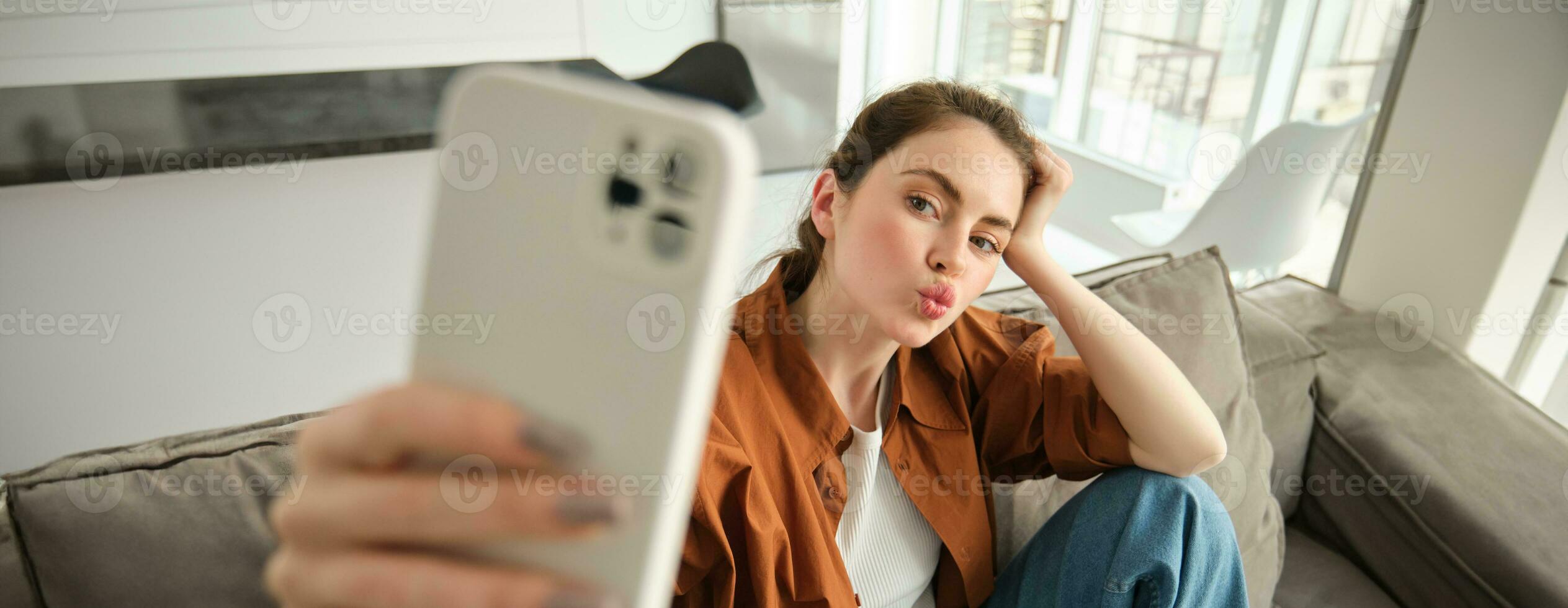 retrato do fofa jovem mulher, moderno menina levando selfies em Smartphone aplicativo, posando em sofá, estende dela branco Móvel telefone para levar foto, franzir lábios para foto