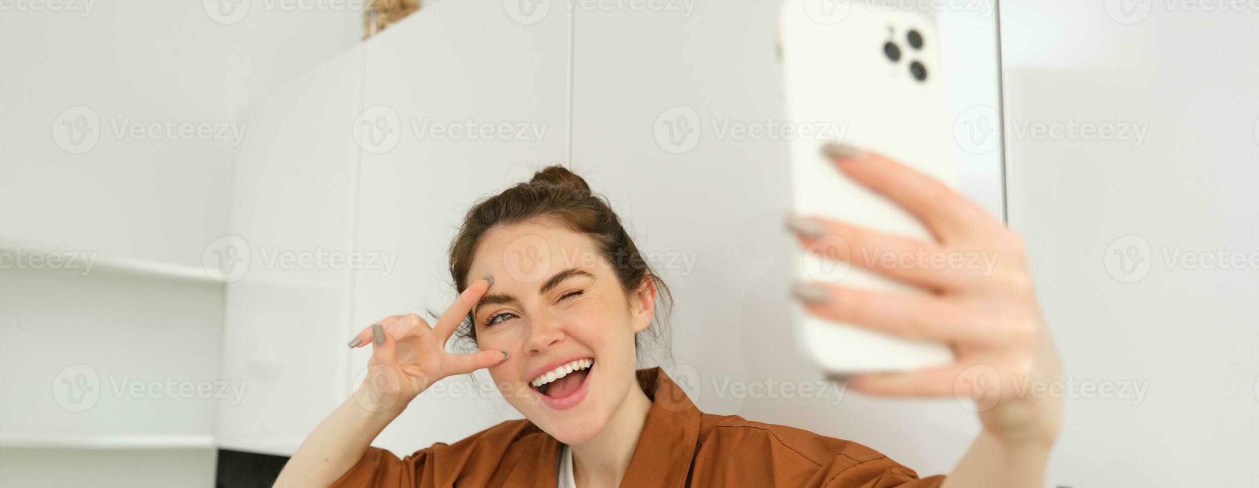 fechar acima retrato do positivo, feliz jovem mulher levando selfie em Móvel telefone, posando para foto em Smartphone, mostrando Paz placa e sorridente às Telefone Câmera