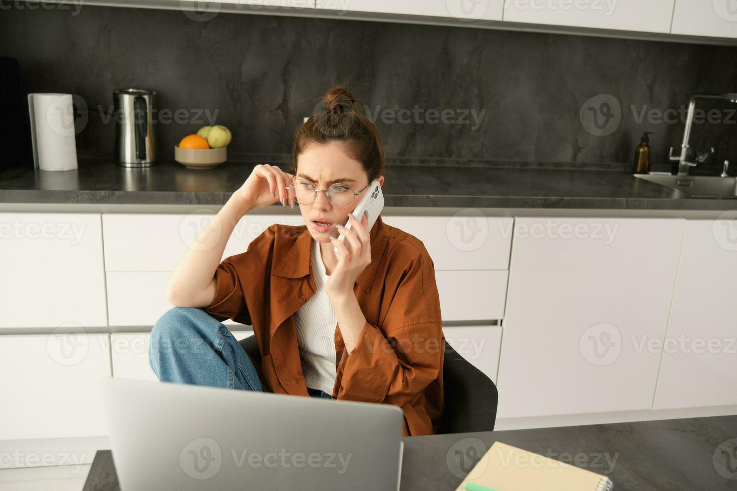 retrato do ocupado jovem trabalhadores por conta própria mulher, trabalhando a partir de lar, tem escritório dentro dela plano, sentado perto computador portátil, respostas telefone chamar, falando com sério, preocupado face expressão foto