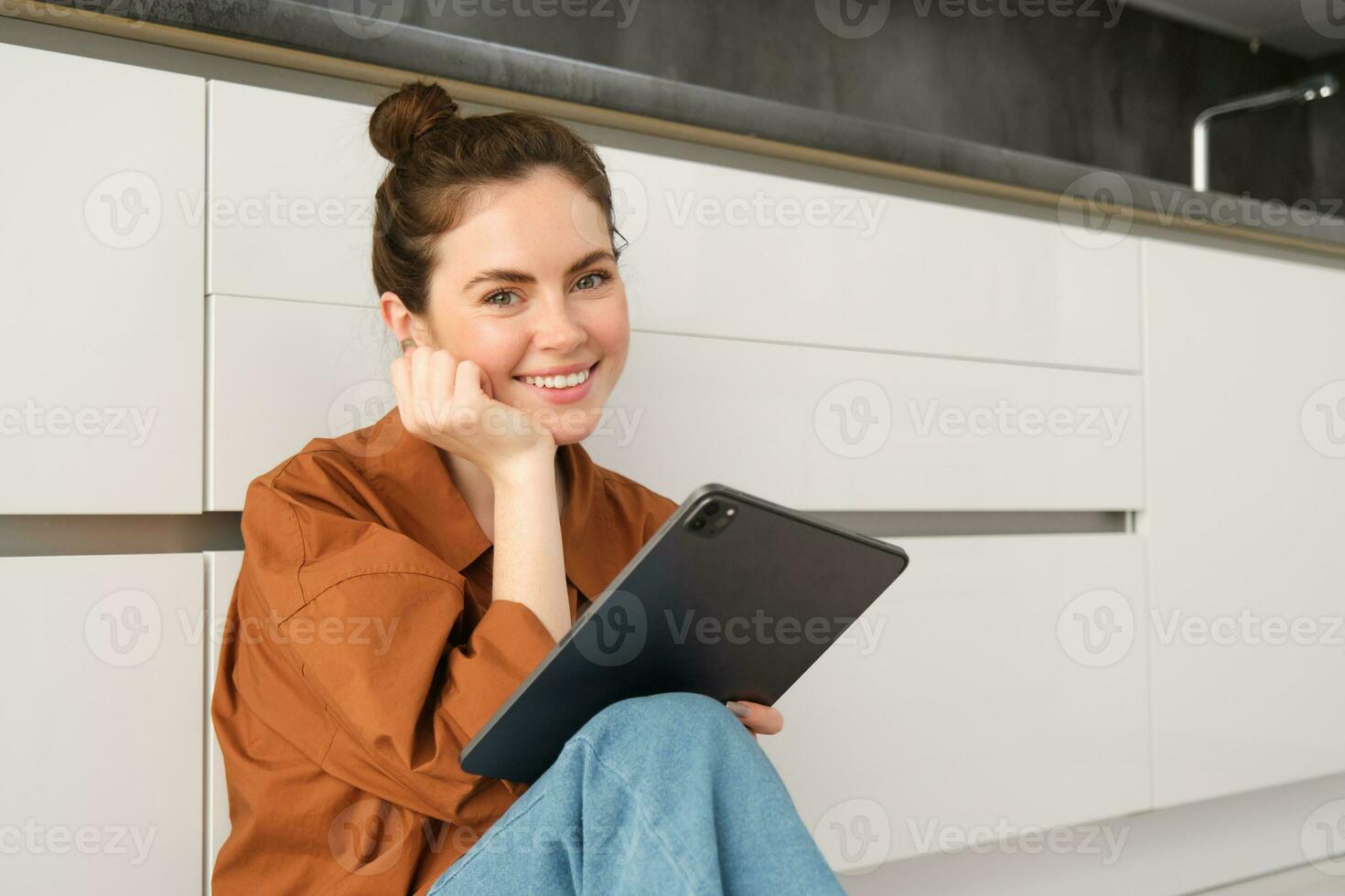 retrato do jovem lindo mulher sentado em cozinha chão com digital tábua, navegando notícia alimentar, social meios de comunicação aplicativo em engenhoca, sorridente e olhando feliz foto