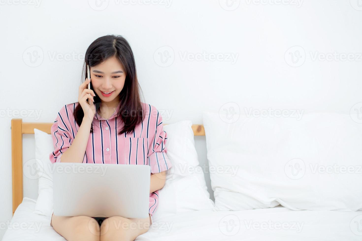 jovem mulher asiática sentada na cama, usando o computador laptop e falando com um telefone inteligente. foto