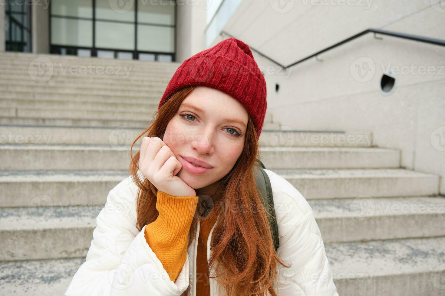 fechar acima retrato do lindo ruiva menina dentro vermelho chapéu, urbano mulher com sardas e gengibre cabelo, senta em escadas em rua, sorrisos e parece linda foto