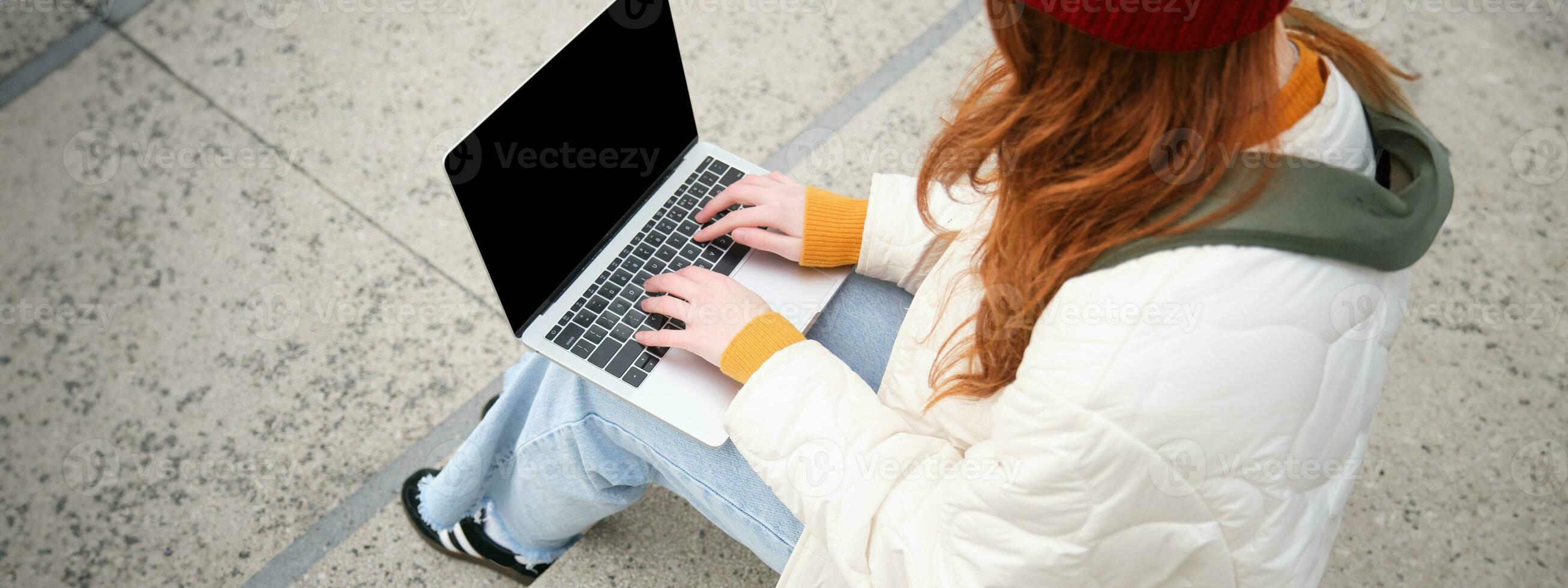 traseiro Visão do menina aluna digitando em computador portátil teclado, em branco Preto tela para local na rede Internet inscrição anúncio, sentado em escadas ao ar livre, conecta para público Wi-fi foto
