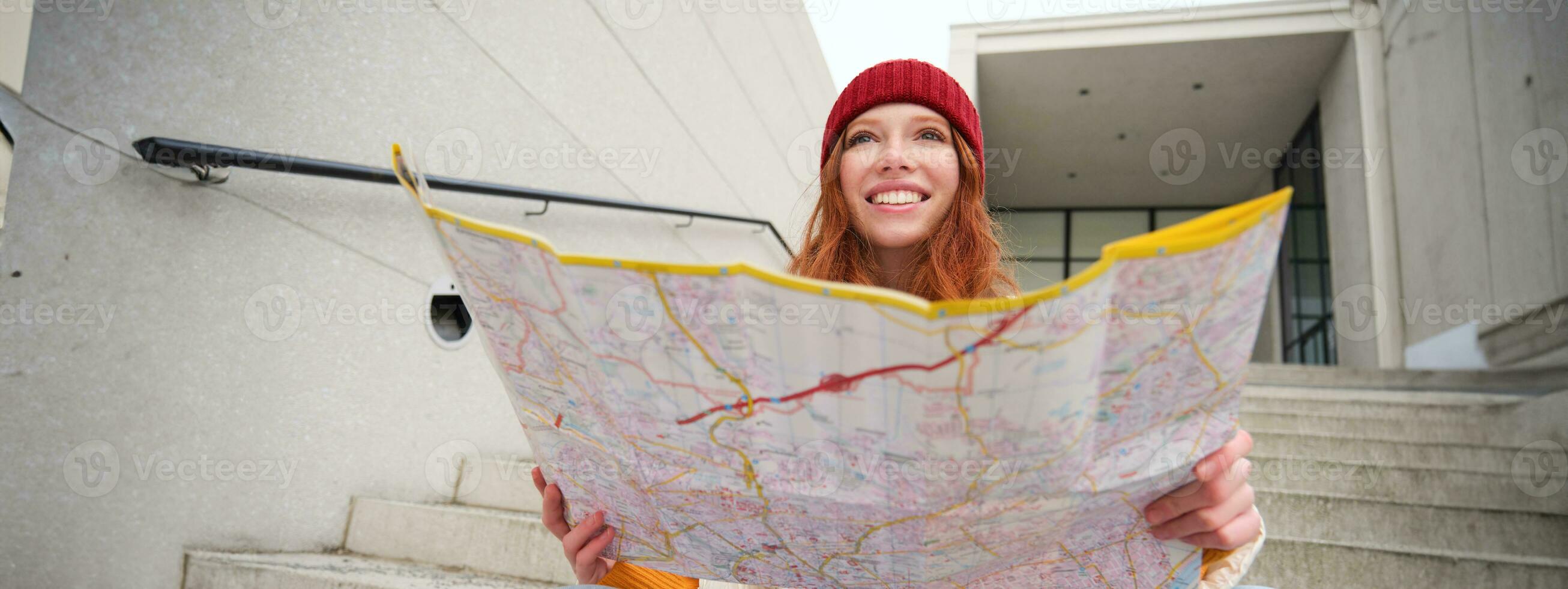 jovem sorridente ruiva garota, turista senta em escadas ao ar livre com cidade papel mapa, olhando para direção, viajante mochileiro explora cidade e parece para passeios turísticos foto