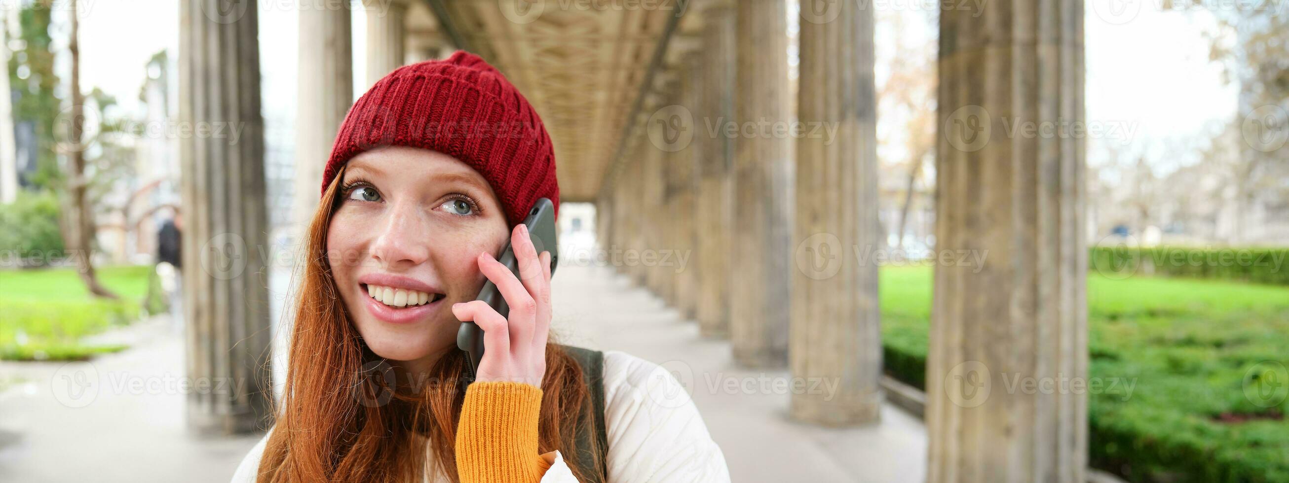 sorridente fofa ruiva mulher faz uma telefone chamar, detém Telefone perto ano, tem Móvel conversação, usando Smartphone em rua foto