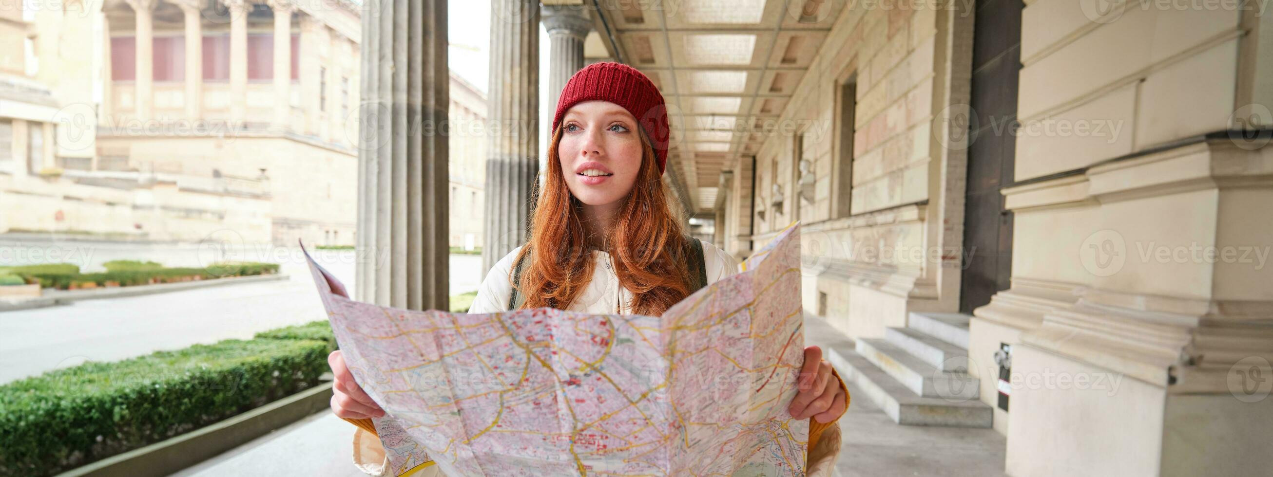 aventureiro ruiva menina anda em dentro Cidade com papel mapa, explora cidade Como turista, parece para popular turismo atrações, parece por aí animado e sorrisos foto