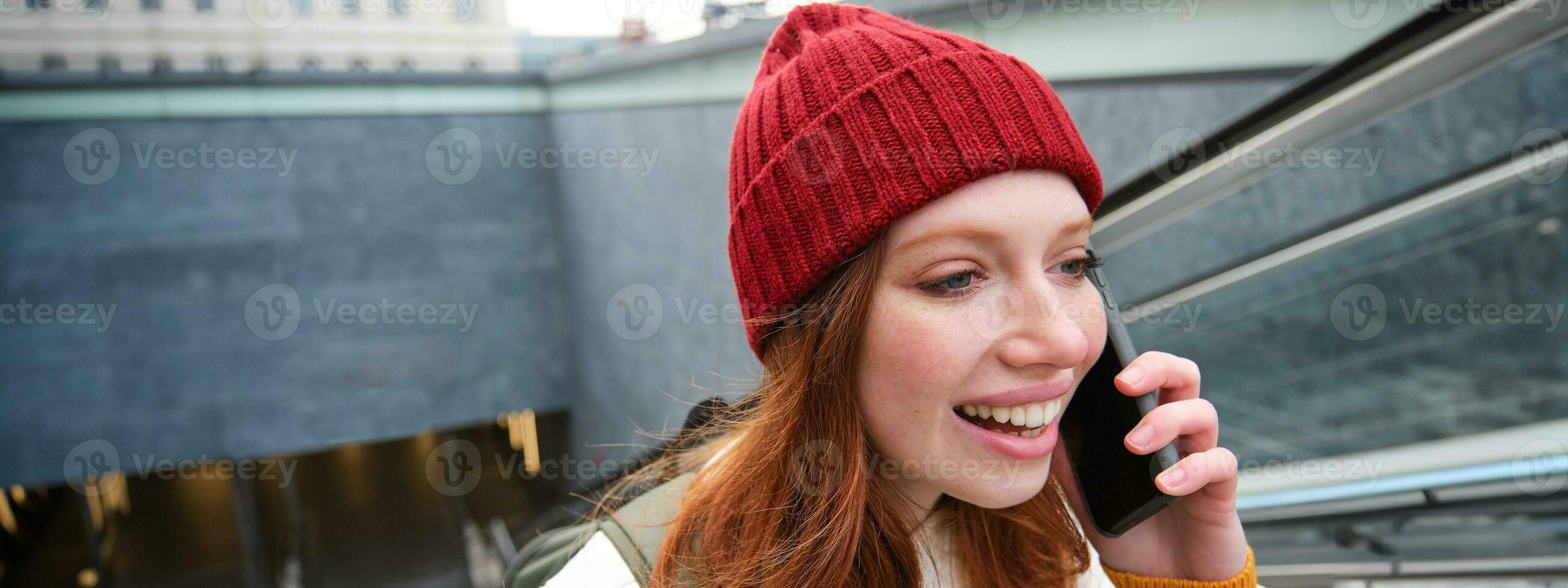 retrato do jovem ruiva mulher anda em por aí cidade, vai acima escadas com Móvel telefone, fala em Smartphone e sorrisos foto