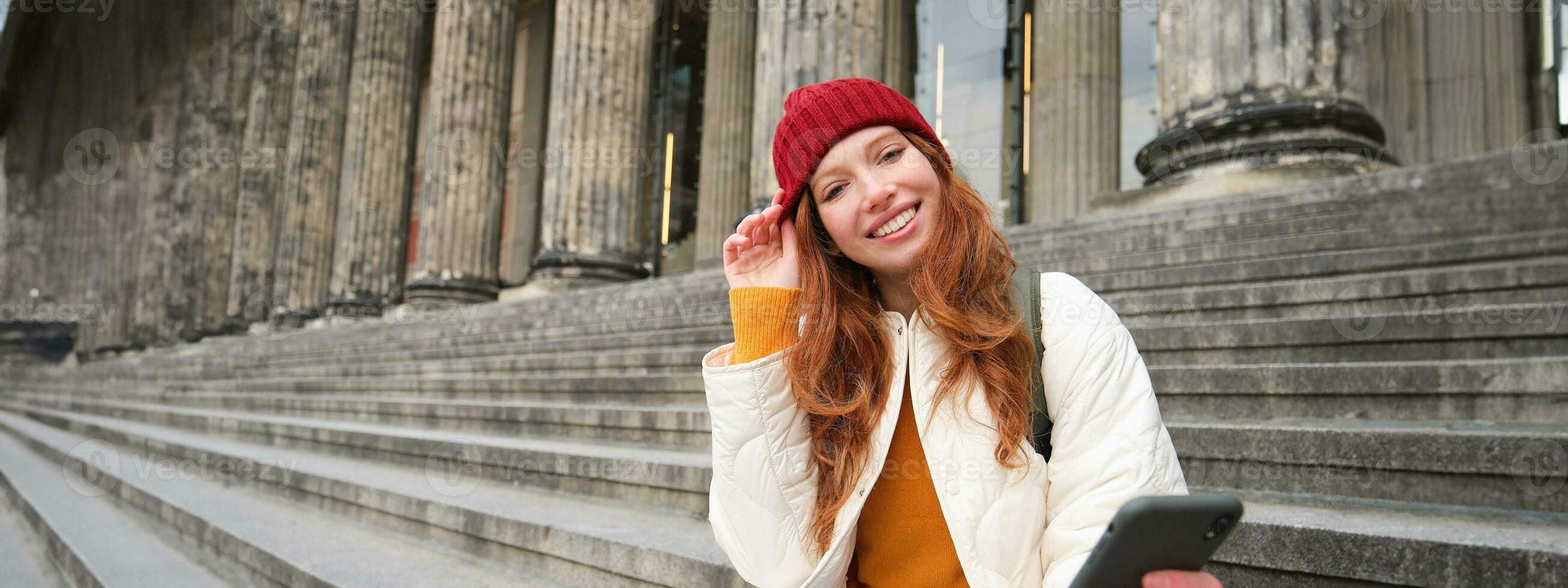 retrato do jovem ruiva mulher dentro vermelho chapéu, sentado em escadaria, turista parece às dela Móvel telefone, descansos em Escadaria do museu, conecta público Wi-fi foto