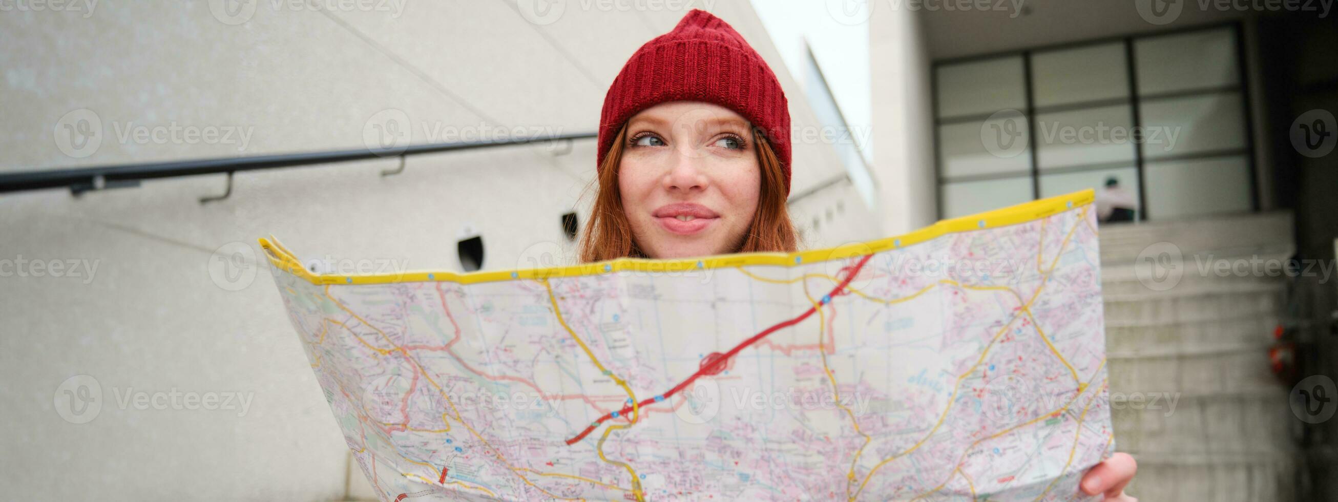 jovem sorridente ruiva garota, turista senta em escadas ao ar livre com cidade papel mapa, olhando para direção, viajante mochileiro explora cidade e parece para passeios turísticos foto