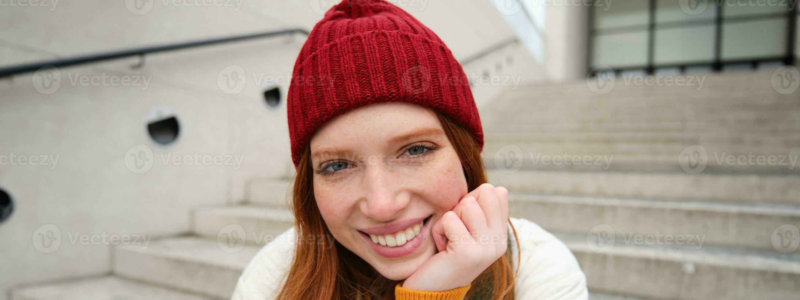 lindo ruiva estudante, menina dentro vermelho chapéu, sorrisos sincero, parece feliz e relaxado, senta em escadas ao ar livre foto