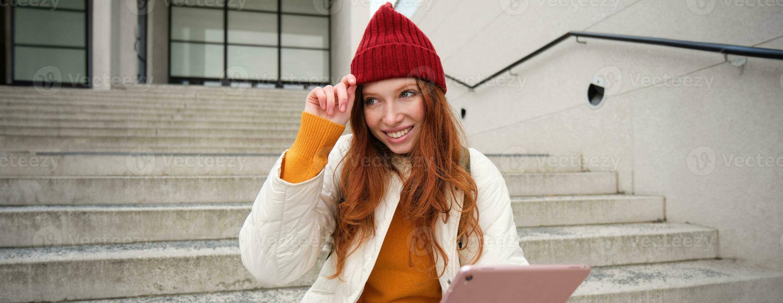 lindo ruiva fêmea modelo, gengibre menina com digital tábua, senta em escadas ao ar livre, lê em dela engenhoca, usa Internet inscrição e Wi-fi em rua foto