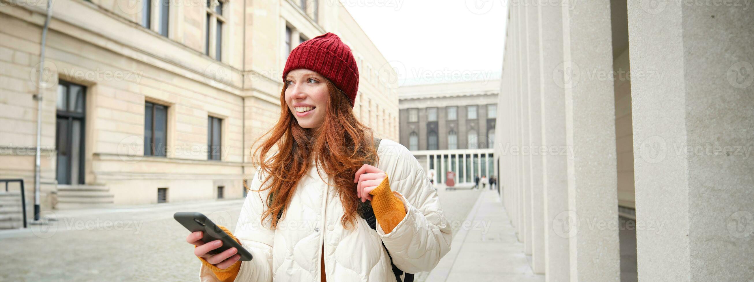 lindo sorridente garota, turista com mochila, segurando Smartphone, usando mapa em Móvel telefone aplicativo, olhando para passeios turísticos dentro Internet aplicativo, em pé ao ar livre foto