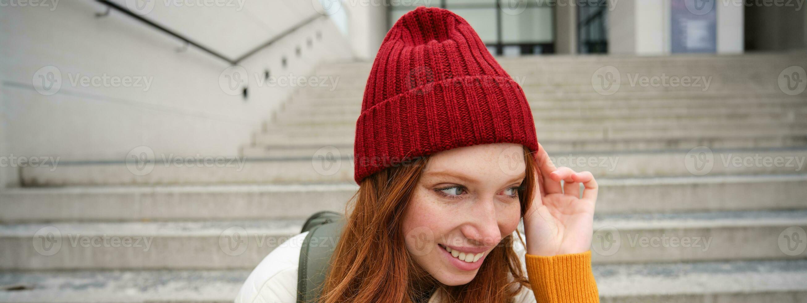 fechar acima retrato do lindo ruiva menina dentro vermelho chapéu, urbano mulher com sardas e gengibre cabelo, senta em escadas em rua, sorrisos e parece linda foto