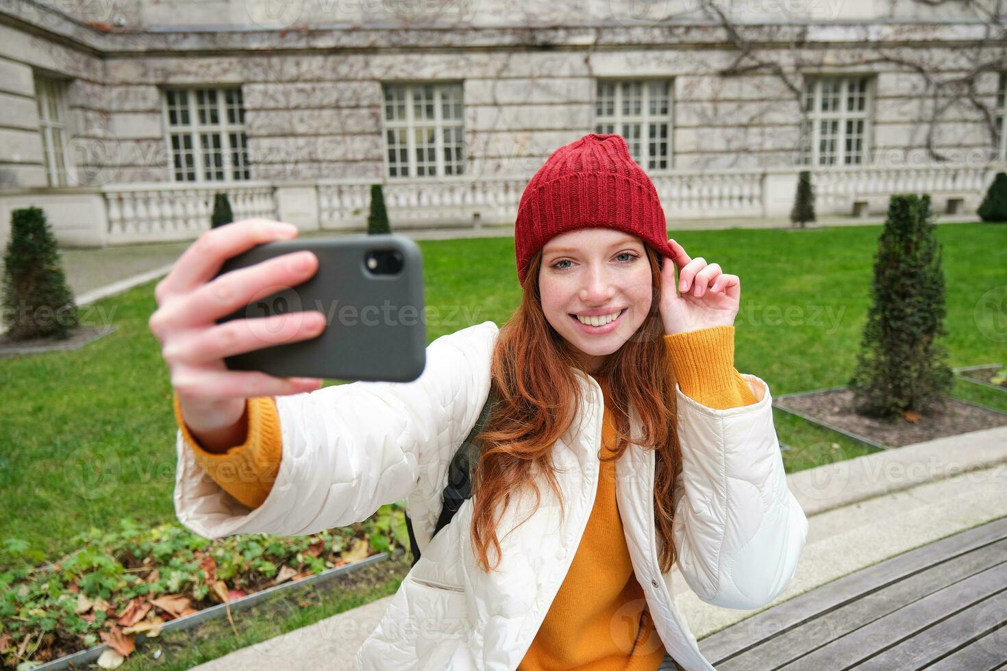 lindo sorridente gengibre menina dentro chapéu, posando para foto em Móvel telefone, leva selfie dentro parque com fofa face expressão